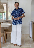 A woman stands in a bright, modern kitchen wearing a navy blue cotton short-sleeve relaxed fit button-up shirt with a white leaf pattern and white wide-leg pants.
