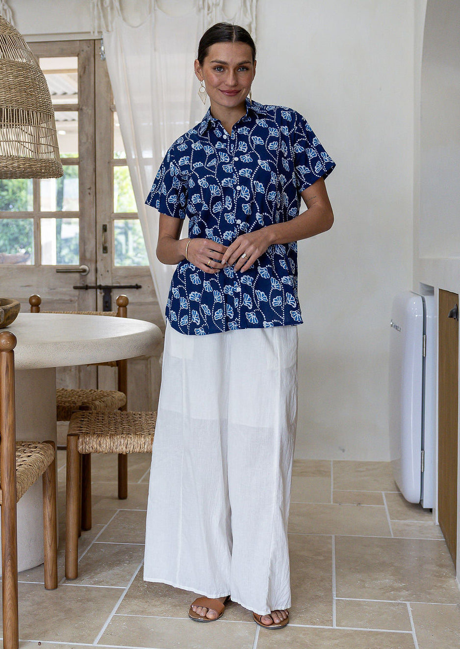 A woman stands in a bright, modern kitchen wearing a navy blue cotton short-sleeve relaxed fit button-up shirt with a white leaf pattern and white wide-leg pants.