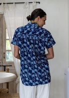 A woman viewed from behind wearing a navy blue cotton short-sleeve shirt with a white leaf block-print pattern, paired with white pants