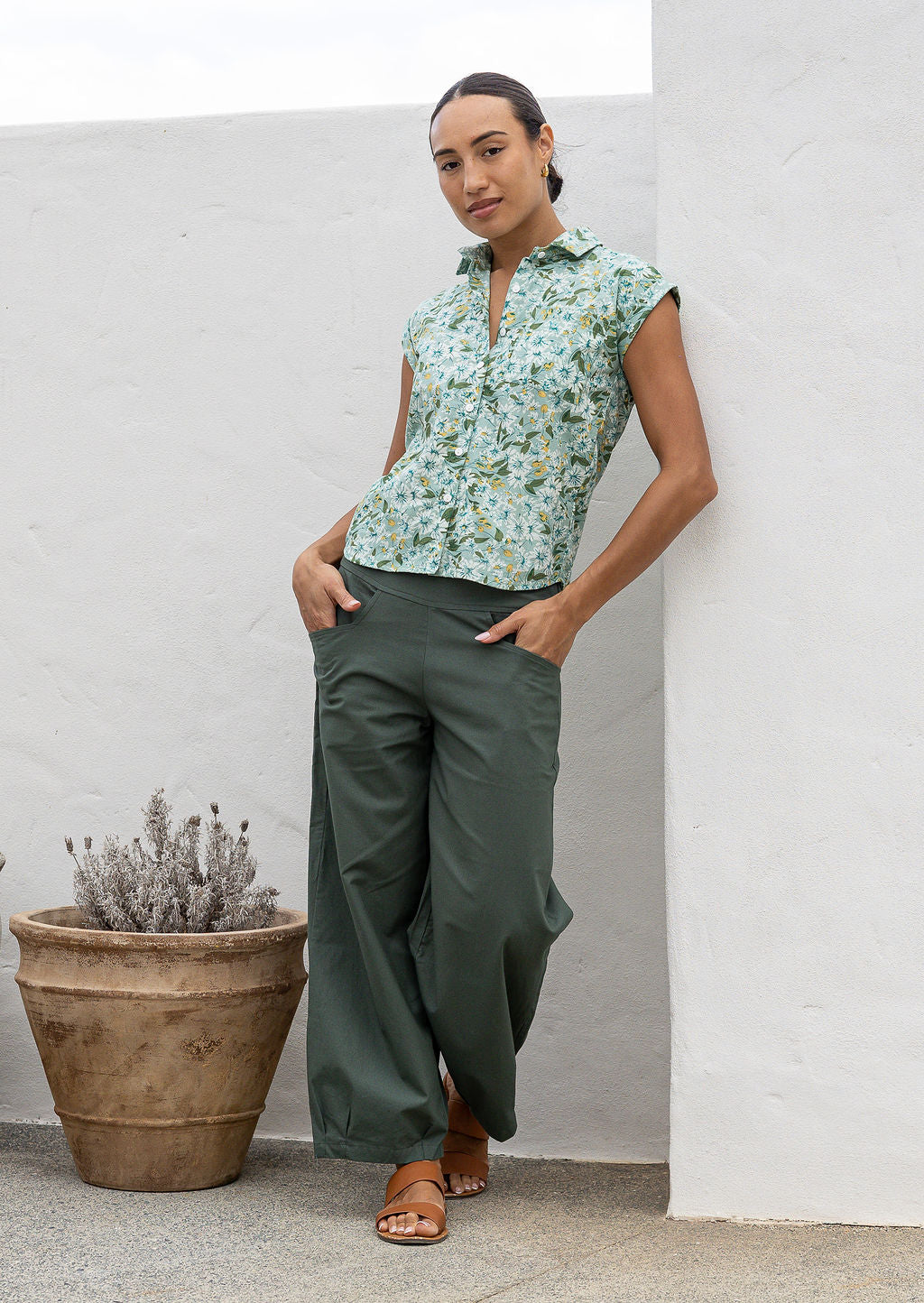 Woman wearing a 100% cotton sage green based floral top standing next to a potted plant against a white wall.