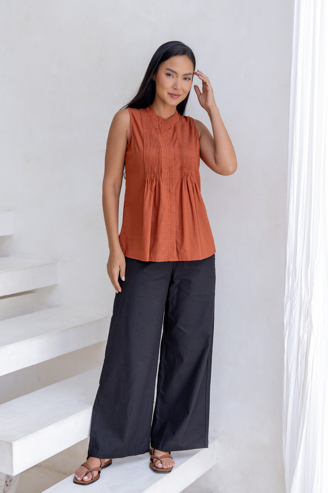 Woman wearing a rust-colored sleeveless top with pleated design, flared fit and high round neckline with button.
