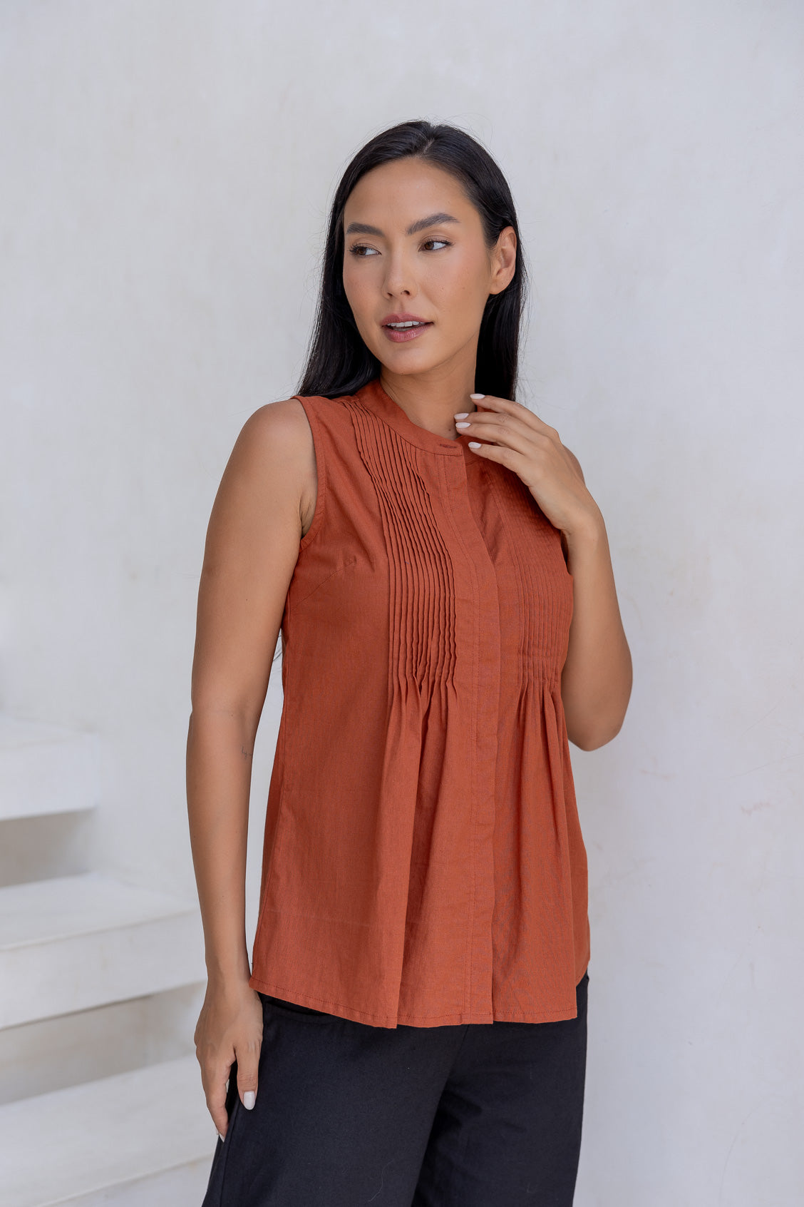 Woman wears without sleeves 100% cotton top in rust shade with mandarin collar, pleated bodice and flared fit below the bust.