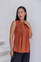 Woman wears without sleeves 100% cotton top in rust shade with mandarin collar, pleated bodice and flared fit below the bust.