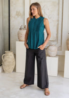 Woman standing with hands in pockets wears a teal sleeveless cotton top and black wide leg pants.