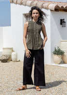 Woman wearing an olive green shade sleeveless cotton blouse with tiny pleats in front and black pants standing in front of a decorative wooden window.