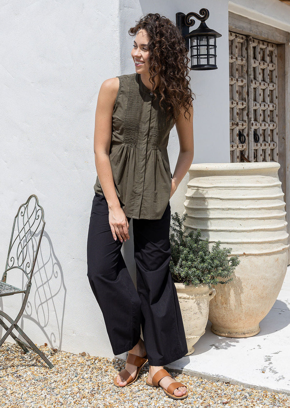 Woman wearing an olive green sleeveless cotton top and black wide-leg pants standing against a white wall.