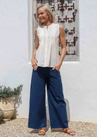 Woman wearing a white sleeveless cotton top and blue wide-leg pants standing against a white wall.