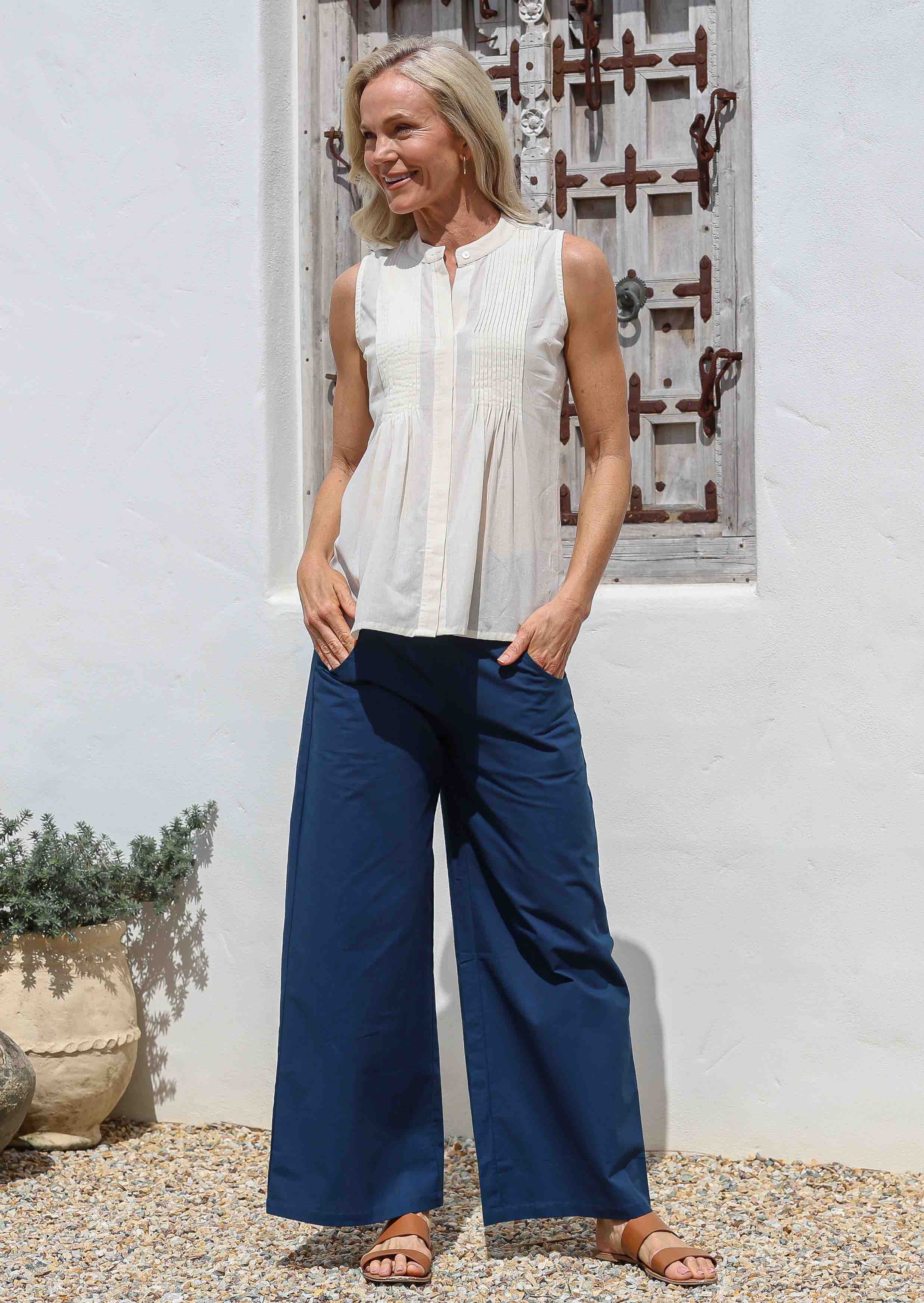 Woman wearing a white sleeveless cotton top and blue wide-leg pants standing against a white wall.