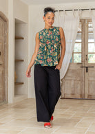 Woman wearing a floral sleeveless top with decorative pleats, high round neckline and black pants in a room setting.