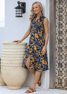 Woman standing next to a large pot wears 100% cotton sleeveless retro black dress with yellow and grey floral print 
