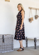Woman standing sideways wearing a collared, sleeveless 100% cotton dress with button-up front in black based floral print