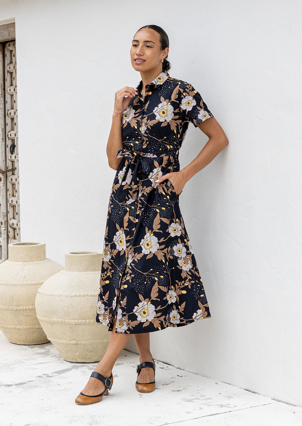 Model leaning on the wall wears 1950s inspired, button through shirt style dress with collar, short sleeves, waist tie and pockets.
