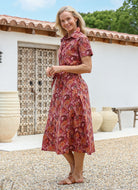 Woman standing sideways wears a retro cotton shirtstyle dress in a Japanese floral print, in front of a rustic wooden door.