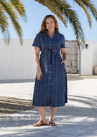 Woman wearing 100 percent cotton button through shirt dress with collar and short sleeves, in a navy based white striped print. 