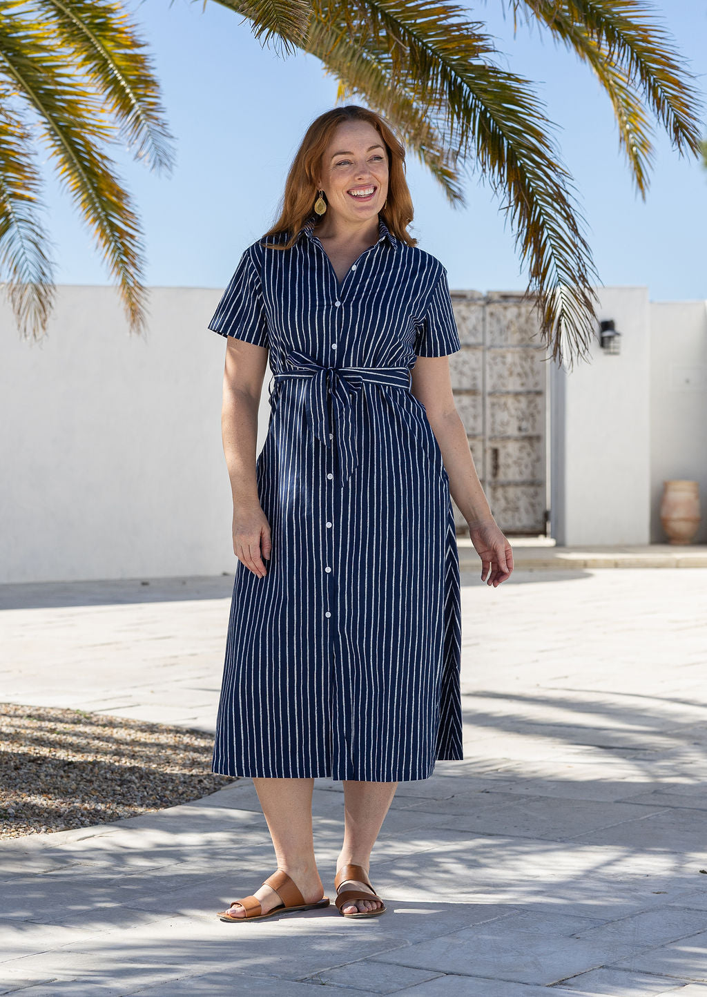 Model poses wearing a shirt style button through cotton dress with collar and a removable waist tie.