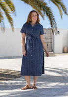 Model poses wearing a shirt style button through cotton dress with collar and a removable waist tie.