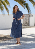 A woman wearing a navy blue based white striped print midi shirt 100% cotton dress with a tie waist, standing outdoors. 