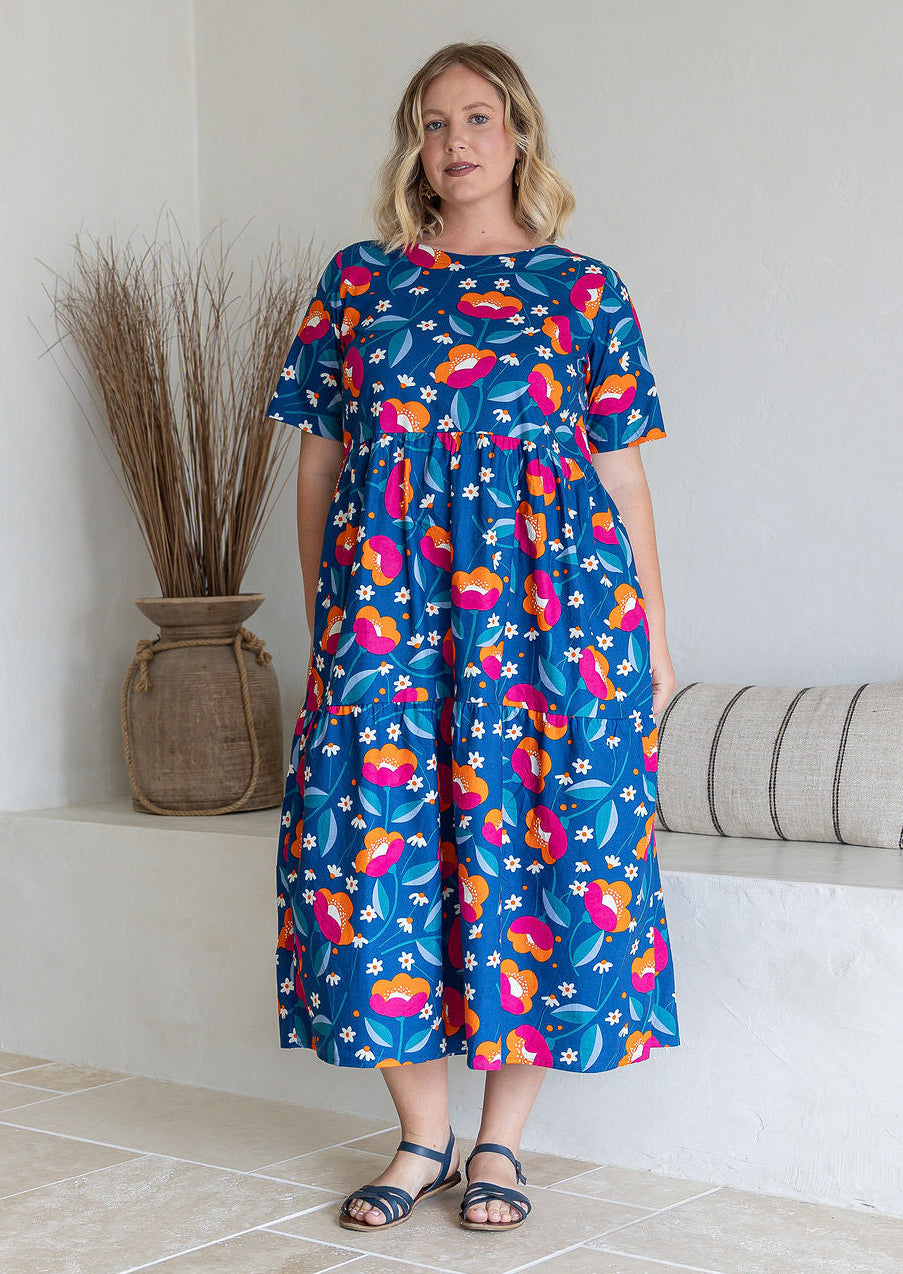Woman wearing a 100% cotton, mid-length royal blue based dress with floral print featuring three tiered design, short sleeves, high round neckline and pockets