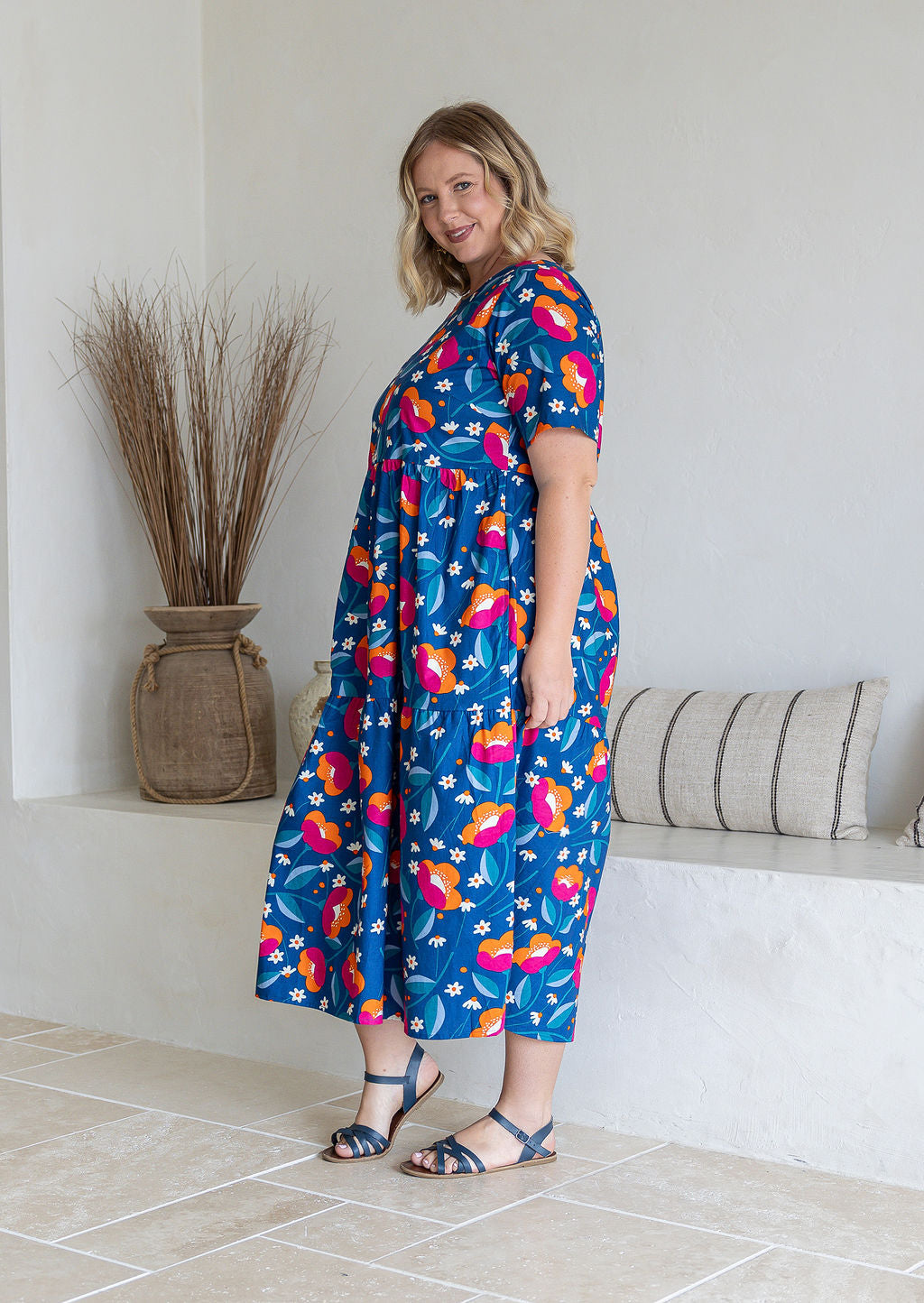 Model stands sideways wearing a floral print, relaxed fit cotton dress with tiers, pockets, and short sleeves, standing indoors.