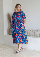 Woman standing wears a floral print cotton dress with three tiered design, short sleeves and hidden side pockets