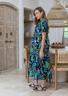 Model stands sideways wearing a floral peacock print, relaxed fit cotton dress with tiers, pockets, and short sleeves, standing in a kitchen. 