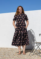 Woman wearing a 100% cotton, mid-length black based dress with floral print featuring three tiered design, short sleeves, high round neckline and pockets