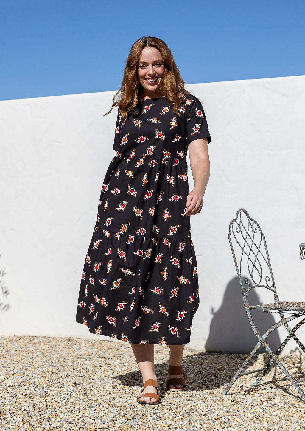 Model wearing a floral Remnant print, relaxed fit cotton dress with tiers, pockets, and short sleeves, standing against a white wall.