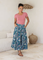 Woman wearing a pink top and green floral cotton floral maxi skirt in a room with a white couch and woven basket.
