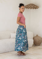 Side view of a Woman wearing a pink top and green floral cotton floral maxi skirt in a room with a white couch and woven basket.