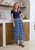 Woman stands wearing a navy top and floral print, shin length cotton skirt with button through front and box pleats.
