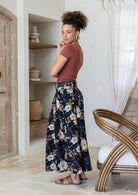 Woman stands sideways wearing a pink top and black floral print, shin length cotton skirt with button through front and box pleats.