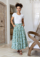 Woman standing with both hand in pockets in a minimalistic room wears 100% cotton button through skirt in sage green based floral print. 