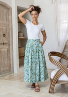 Woman wearing a white top and floral skirt standing in a room with wooden door and chair.