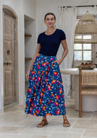 Woman stands wearing a floral print, shin length cotton skirt with button through front and box pleats.