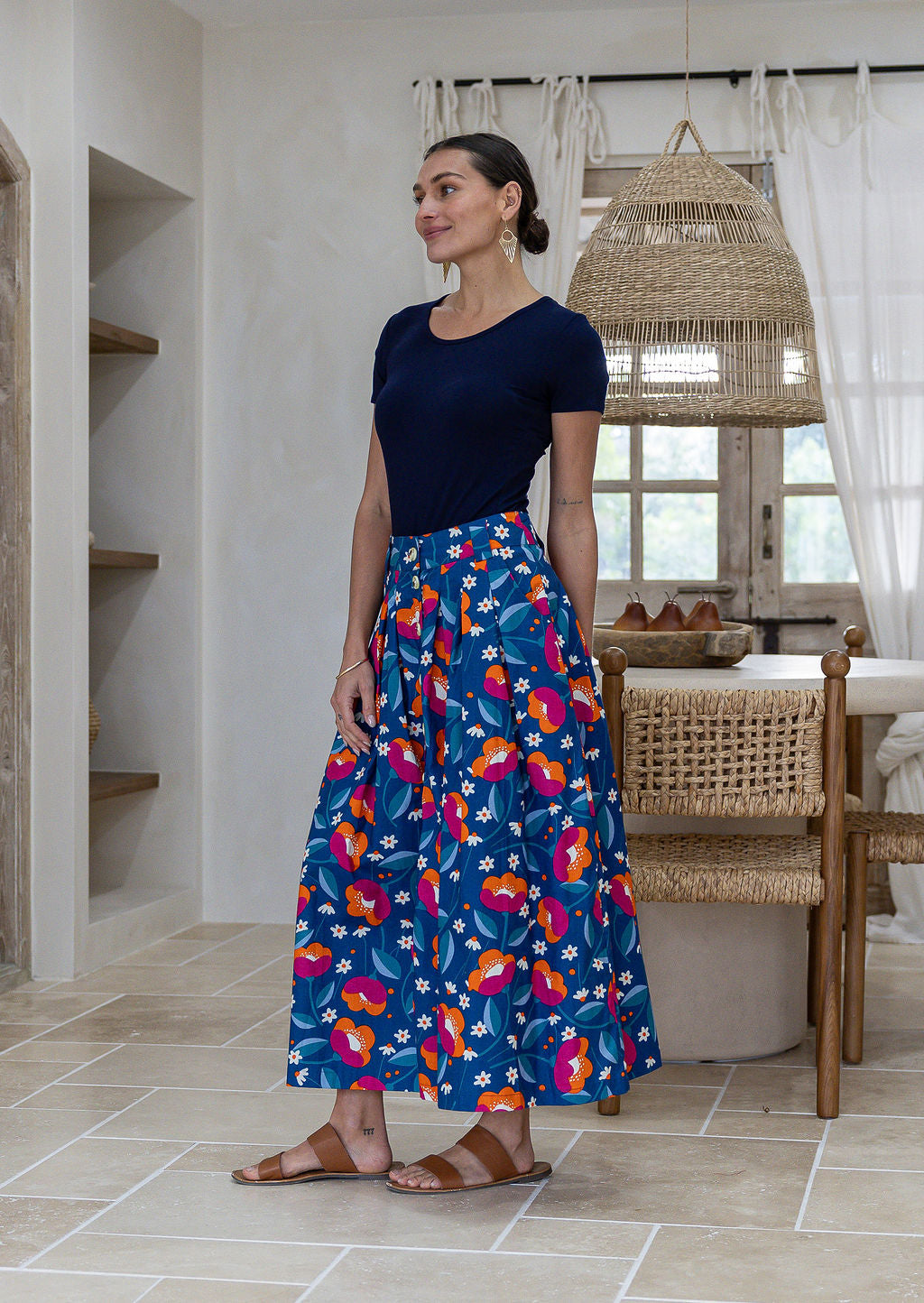Woman stands sideways wearing a basic top and floral print, shin length cotton skirt with button through front and box pleats.