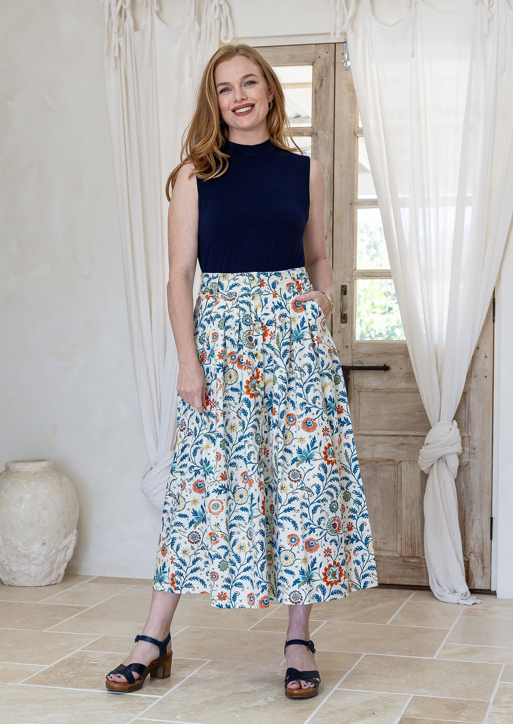 Woman stands wearing a navy top and floral print, shin length cotton skirt with button through front and box pleats.