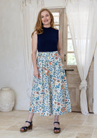 Woman stands wearing a navy top and floral print, shin length cotton skirt with button through front and box pleats.