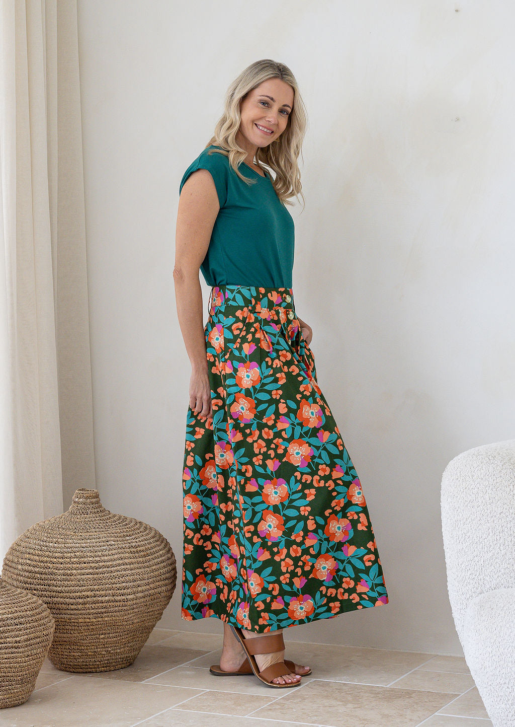 Woman stands sideways wearing a basic top and floral print, shin length cotton skirt with button through front and box pleats.