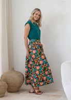 Woman stands sideways wearing a basic top and floral print, shin length cotton skirt with button through front and box pleats.