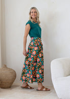 Woman stands wearing a floral print, shin length cotton skirt with button through front and box pleats.
