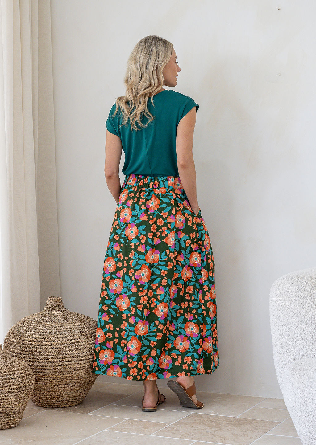 Woman stands backwards wearing a basic top and floral print, shin length cotton skirt with button through front and box pleats.