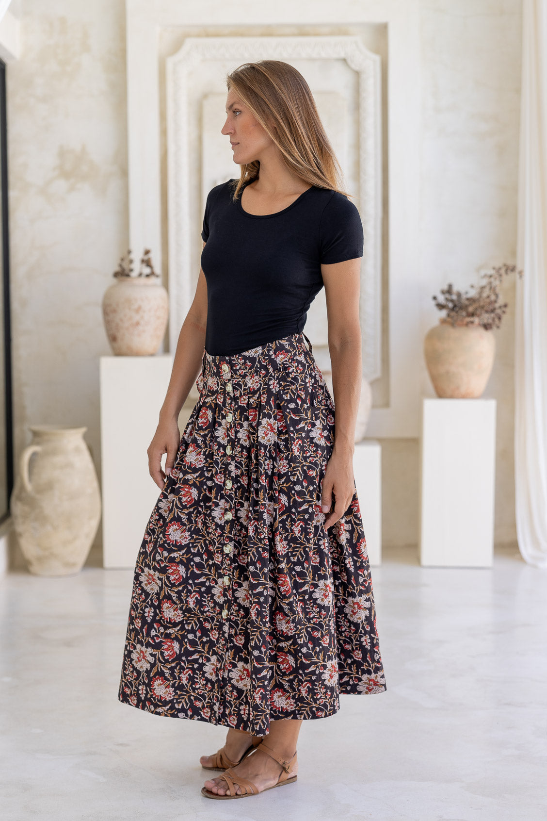 Woman poses in 100% cotton button through a-line skirt with box pleats, belt loops and pockets in black based traditional indian floral print 
