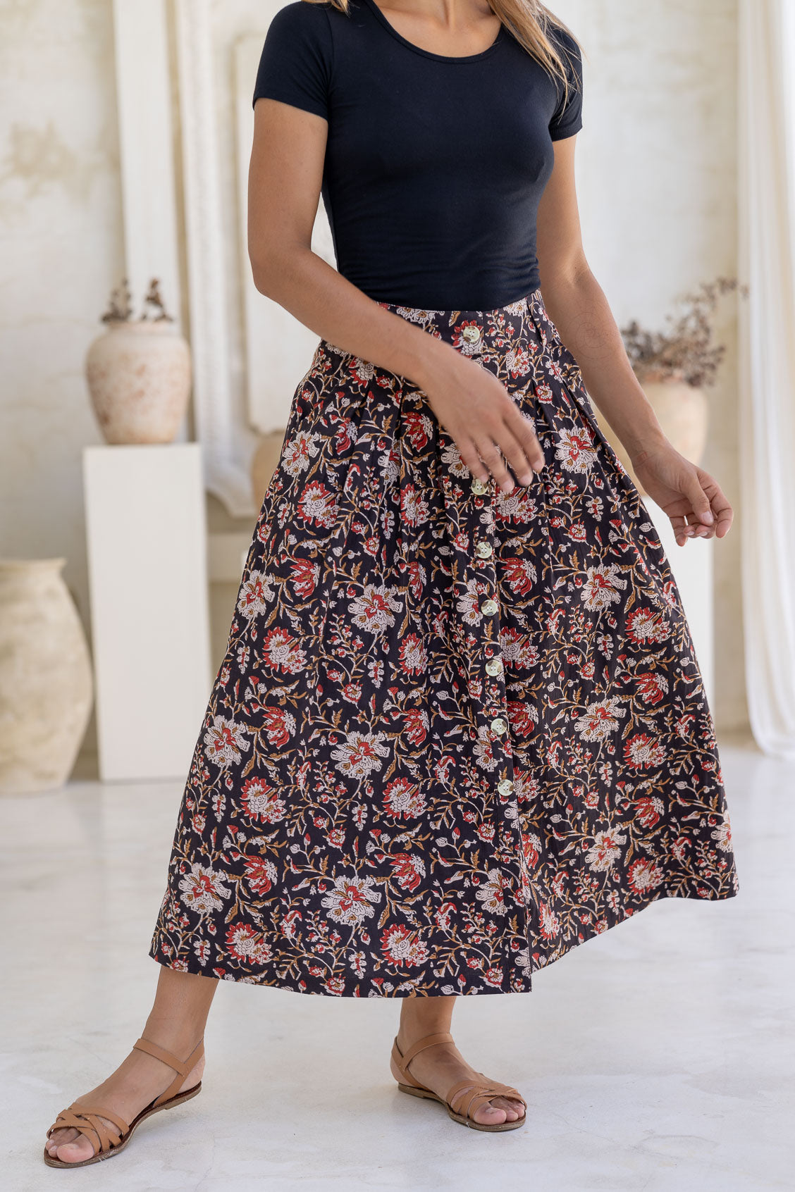 Close up image of a model wearing button through cotton skirt with box pleats and belt loops in black based indian floral print.
