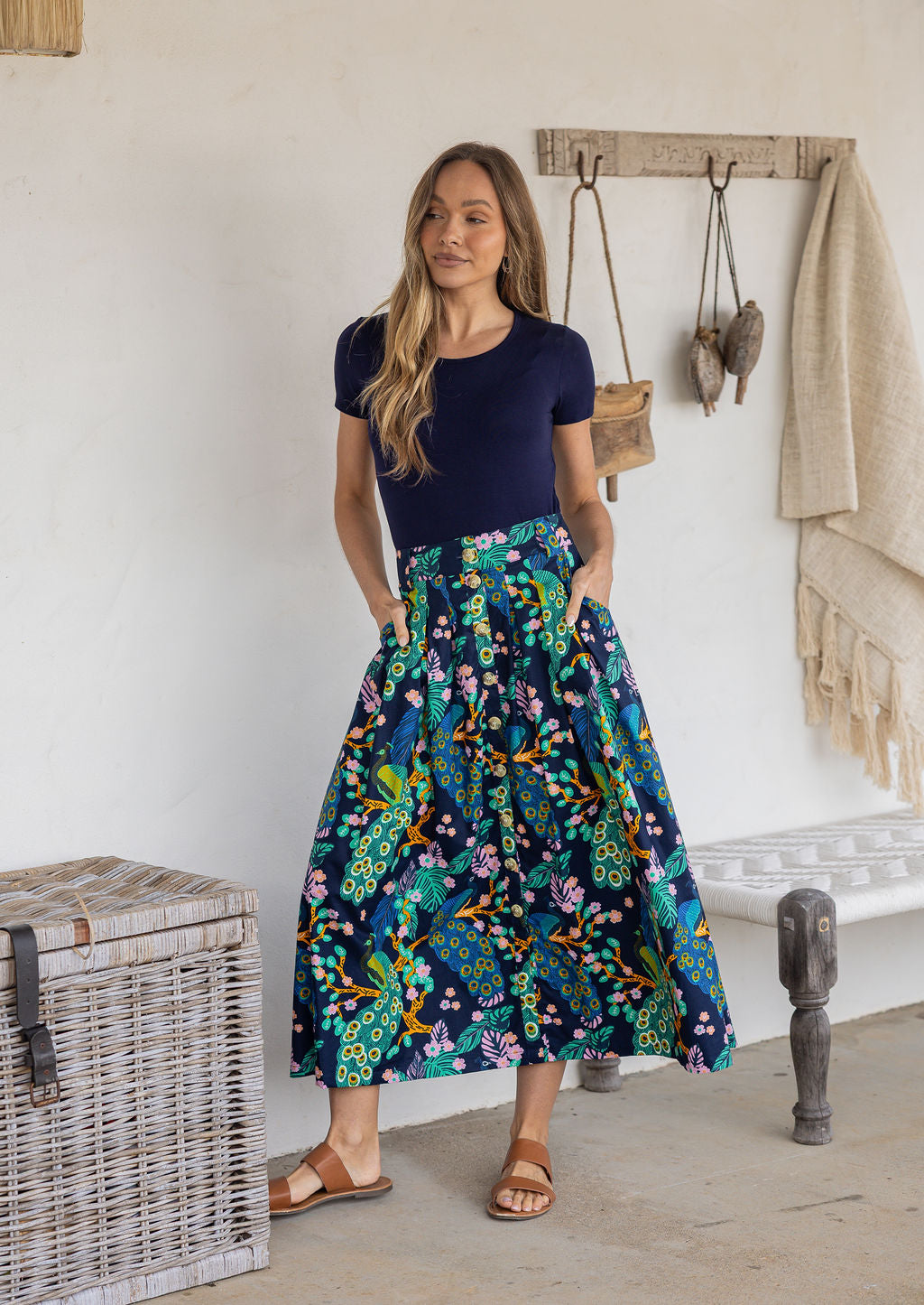 Woman stands wearing a navy top and floral peacock print, shin length cotton skirt with button through front and box pleats.