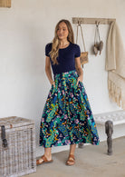 Woman stands wearing a navy top and floral peacock print, shin length cotton skirt with button through front and box pleats.