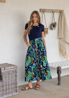 Woman stands wearing a navy top and floral peacock print, shin length cotton skirt with button through front and box pleats.