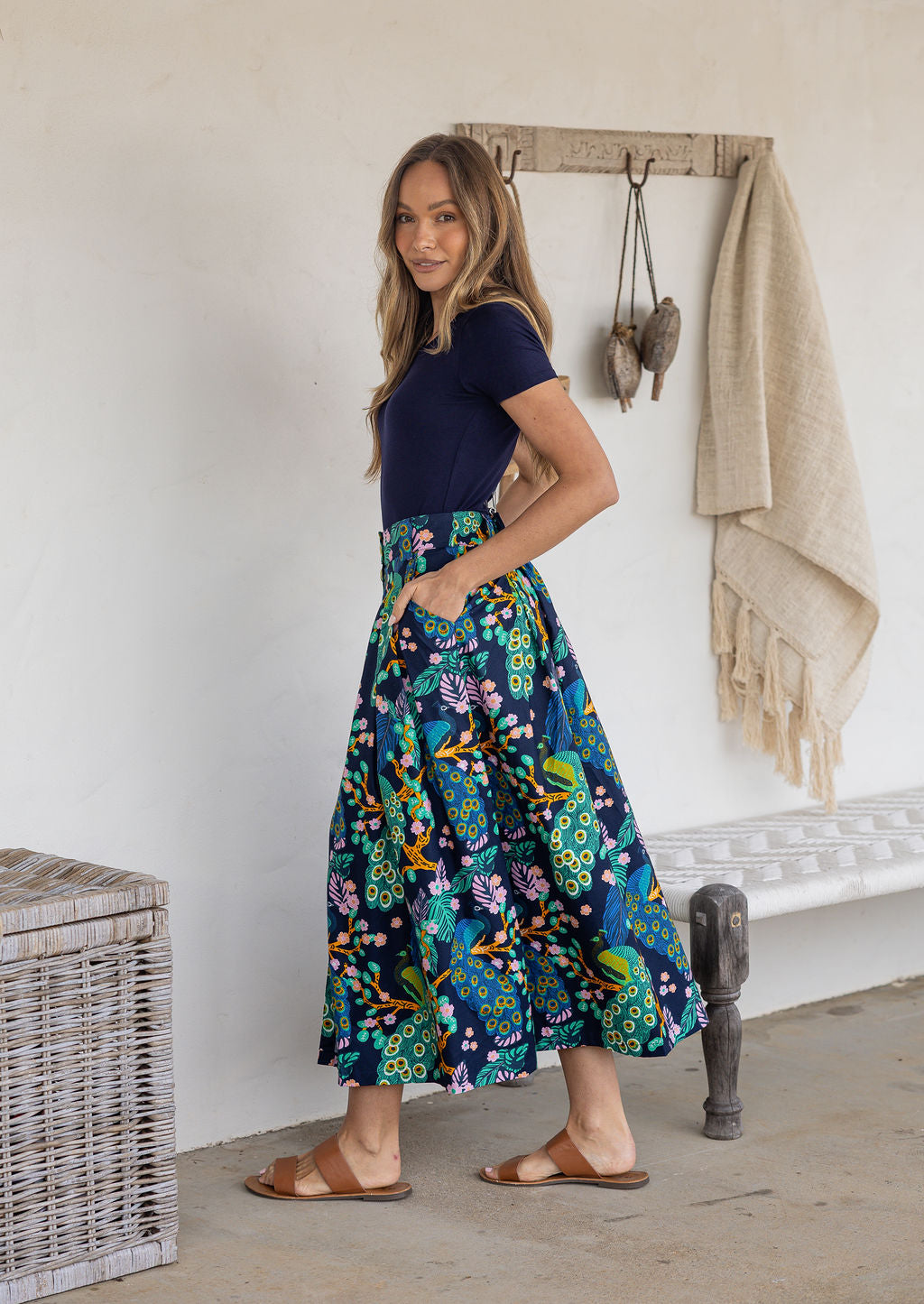 Woman stands sideways wearing a navy top and floral peacock print, shin length cotton skirt with button through front and box pleats.