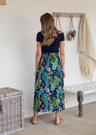 Woman stands backwards wearing a navy top and floral peacock print, shin length cotton skirt with button through front and box pleats.