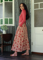 Woman stands sideways wearing a pink top and floral print, shin length cotton skirt with button through front and box pleats.