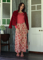 Woman in a red jacket and floral skirt standing in a room with a door and window.
